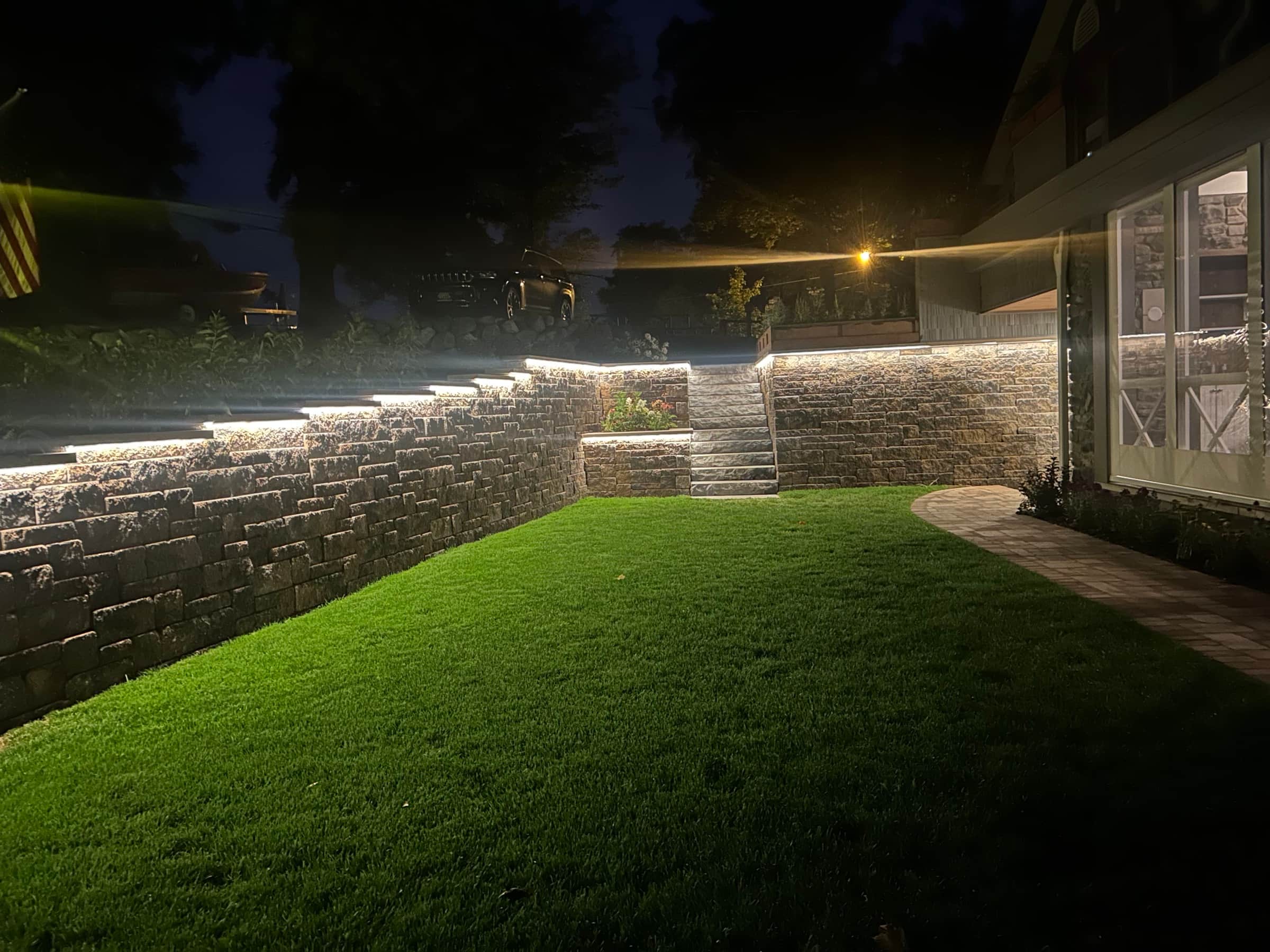 LED-lit stone retaining wall at night with vivid green lawn illuminated by landscape lighting
